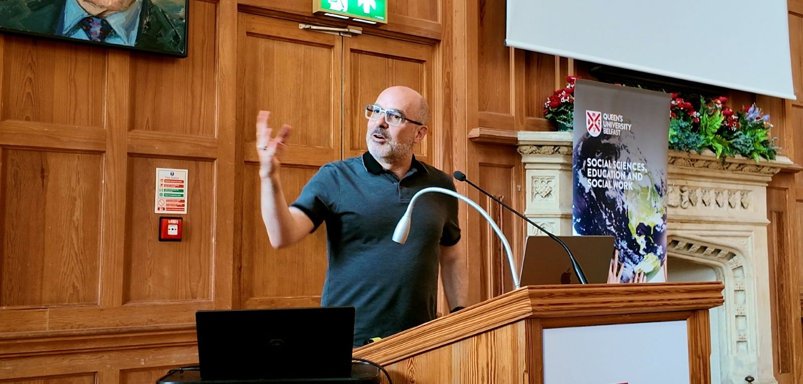 bald man wearing glasses standing a lectern with a wood-panelled wall behind